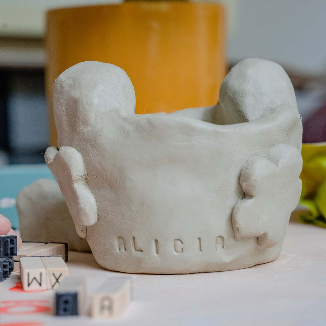 A Sculpd Kids Pottery Kit clay Pinch Pot with cut-out flowers and the name Alicia stamped on it with Sculpd Letter Stamps on a table