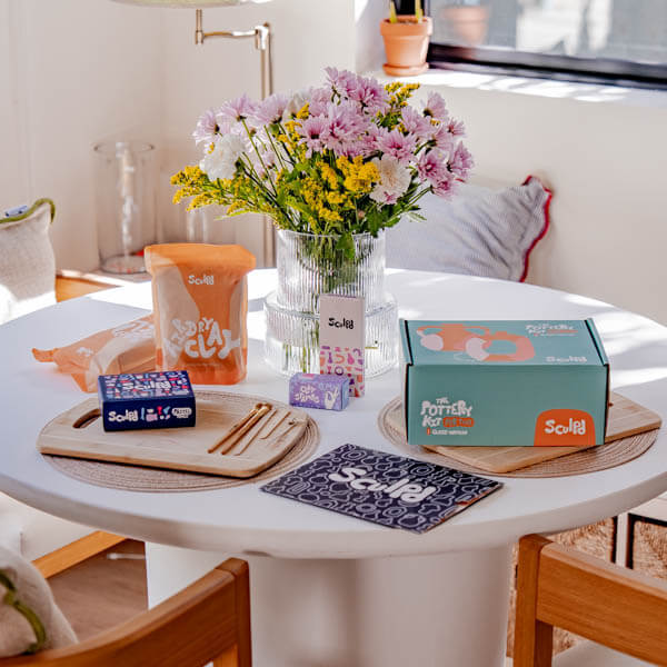 A cozy but fresh-looking setup featuring a Sculpd Pottery Kit and fresh wildflowers in a vase, with sculpting tools arranged, creating a warm, spring ambiance