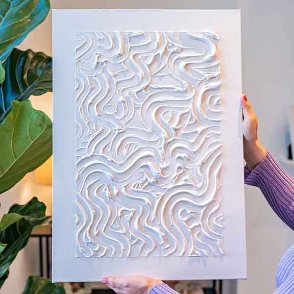 A white textured artwork with a repeating, raised pattern stands on display with greenery in the background made of Sculpd Textured Canvas Kit