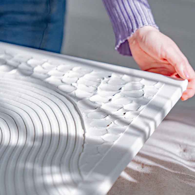 A person holding the edge of a textured canvas artwork featuring intricate, raised patterns made from Sculpd Textured Canvas Kit