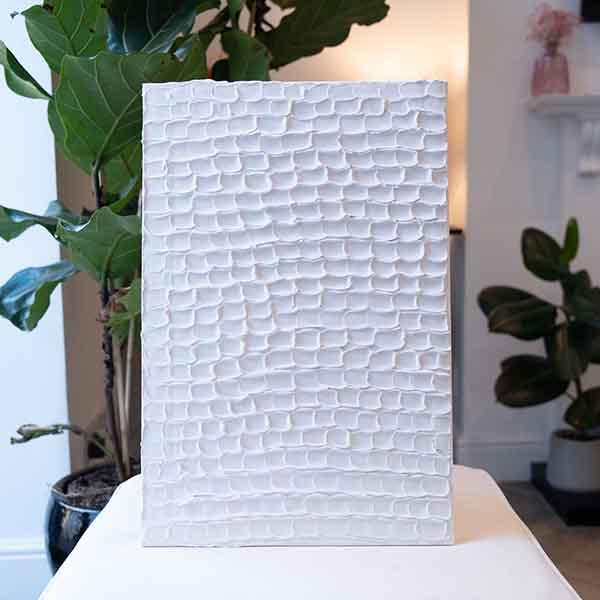 A white textured artwork with a repeating, raised pattern stands on display with greenery in the background made of Sculpd Textured Canvas Kit