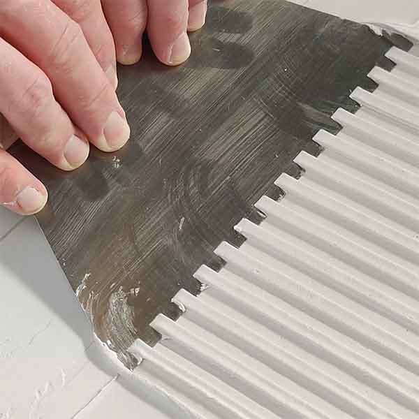 A hand uses a notched scraper to create textured lines in white Textured Paste using Textured Canvas Kit