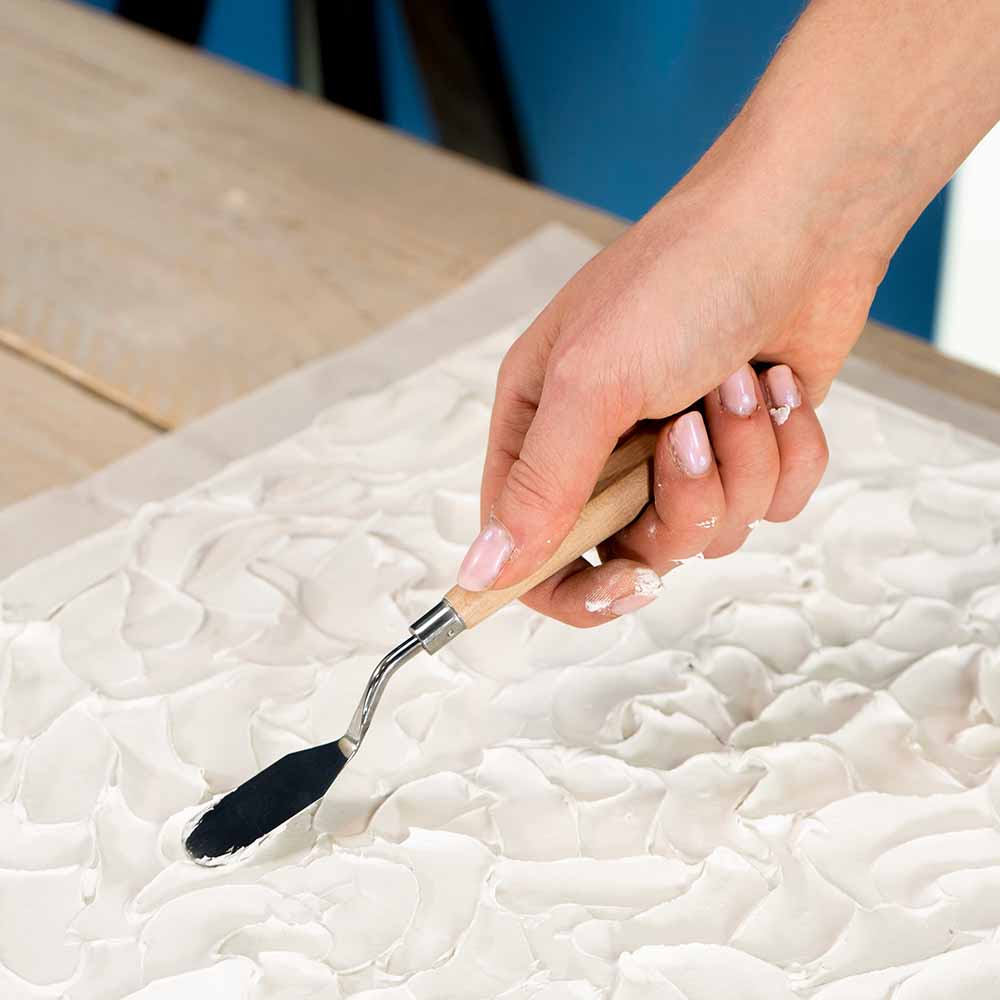 Close-up of a hand using a palette knife to create textured patterns in white Textured Paste using Sculpd Textured Canvas Kit