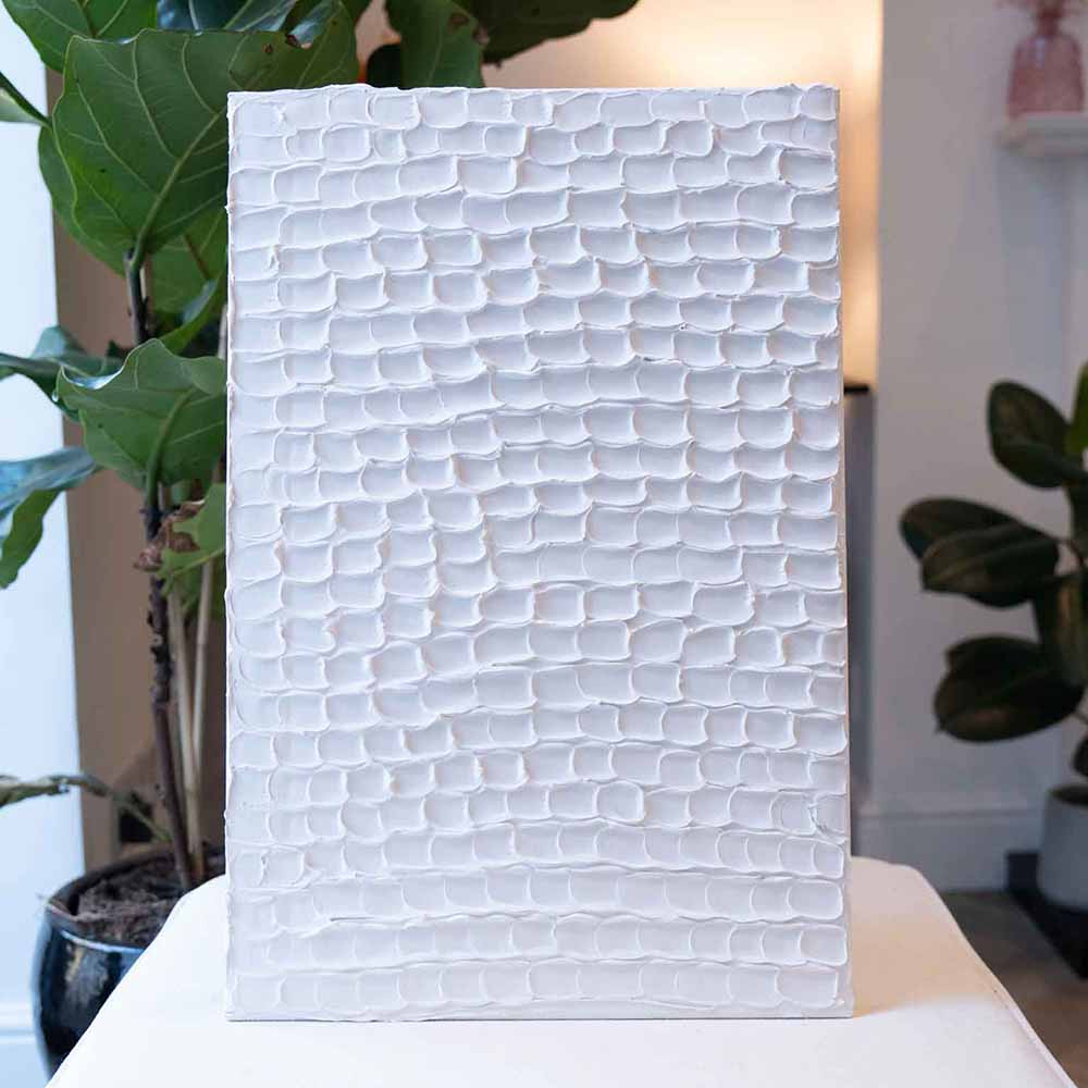 A white textured artwork with a repeating pattern stands on display with greenery in the background made using Sculpd Textured Canvas Kit
