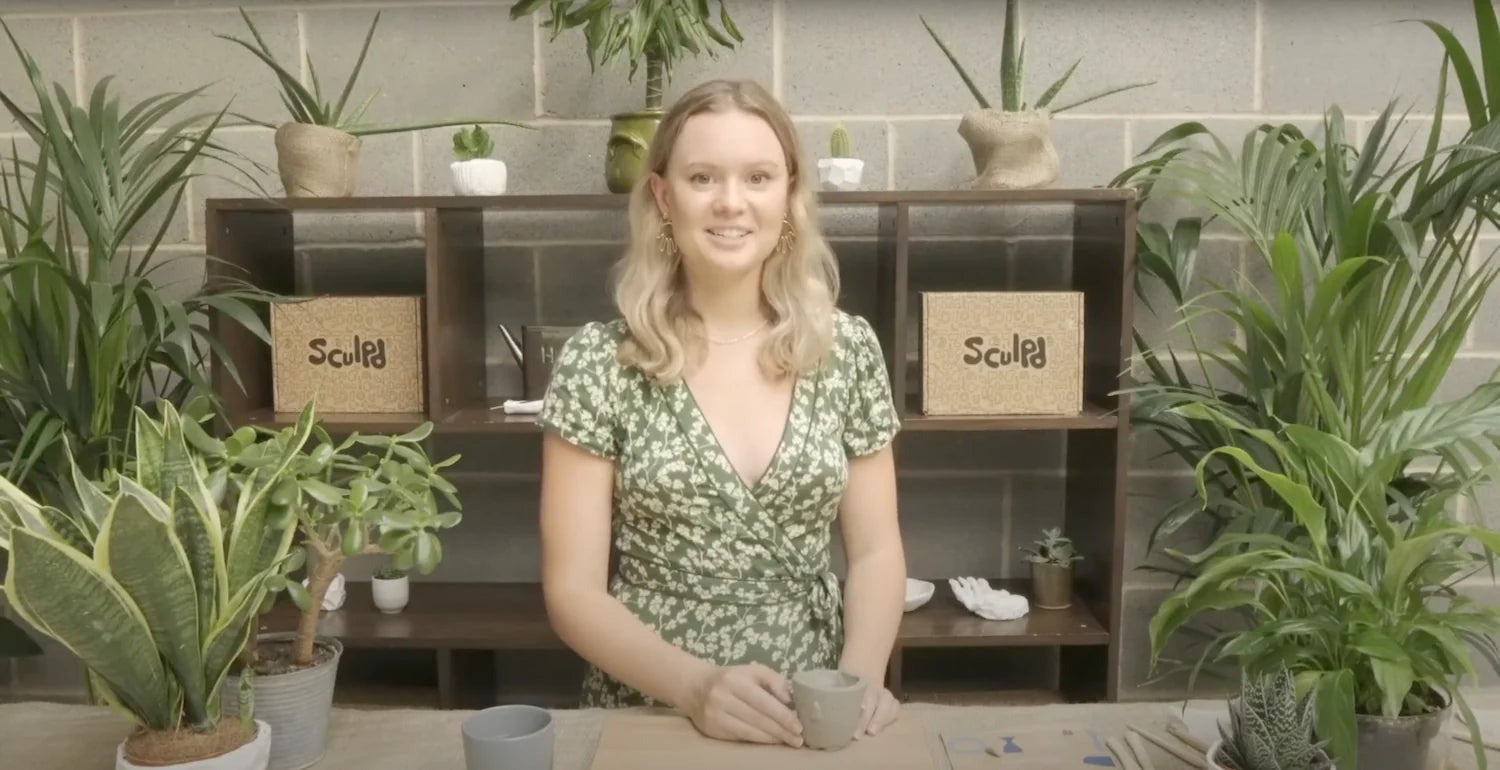 Smiling woman demonstrating pottery techniques, with Sculpd boxes and plants in the background