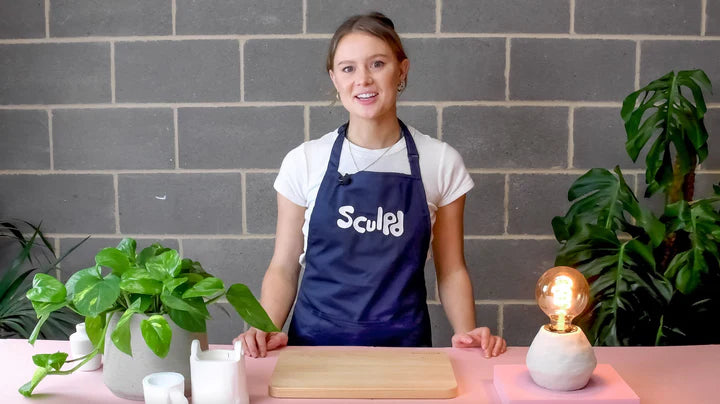 Woman demonstrating pottery techniques with plants, Sculpd tools and a lamp made with the Sculpd Filament Lamp Kit displayed on a table